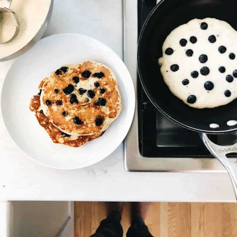 A large white plate of blueberry pancakes with butter melting over them as some cook in a skillet next to it.