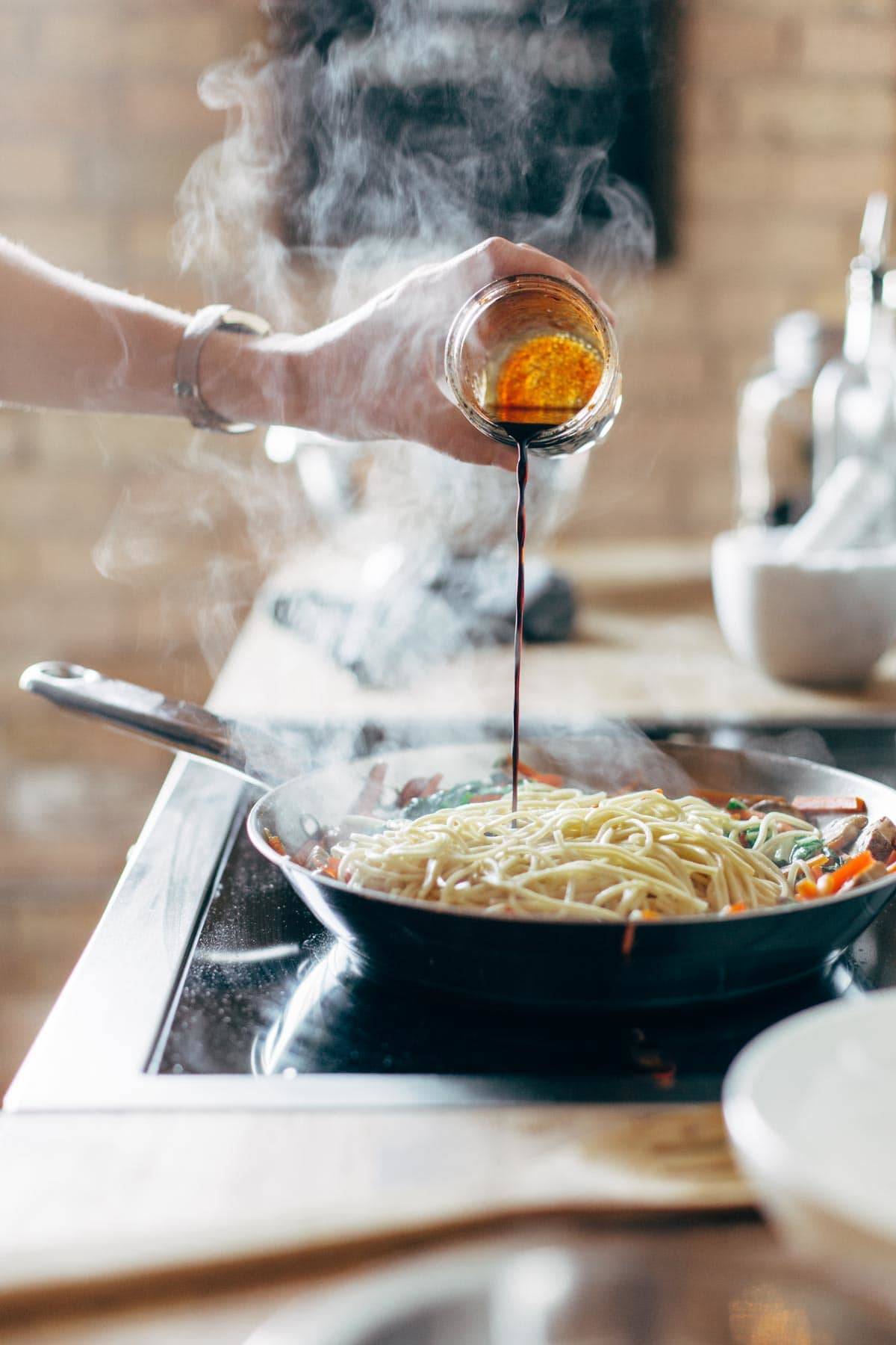 White hand pouring soy sauce on noodles in pan. 