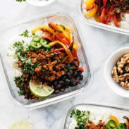 Jalapeno peppers, vegan taco meat, black beans, lime wedges, and red and orange bell peppers in a clear bowl over rice.